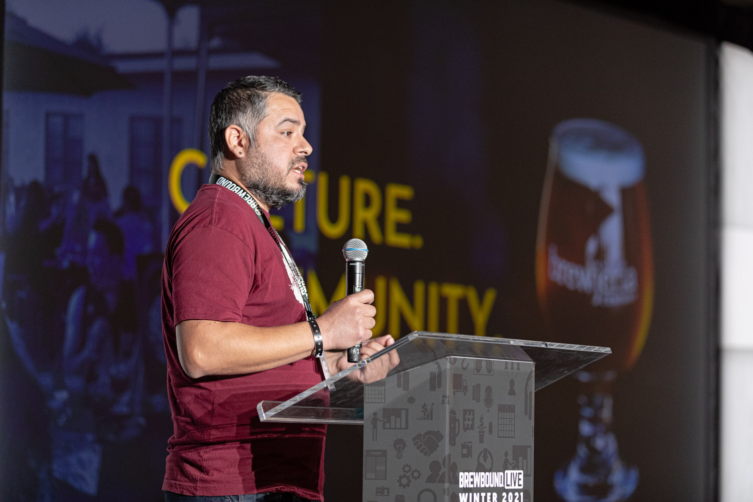 A man in a maroon shirt speaks at the podium in front of a slide with the words Culture and Community.