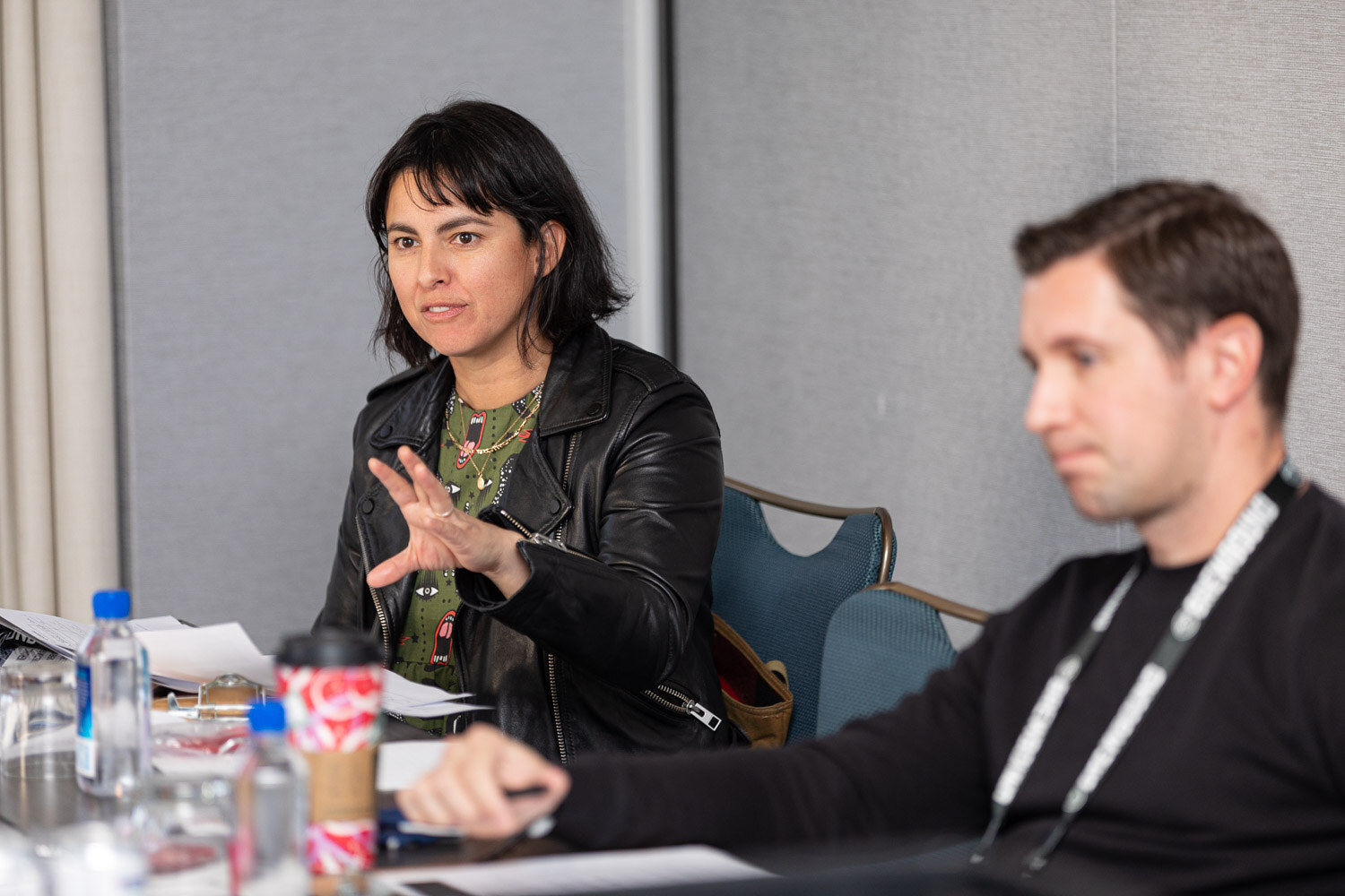 A woman in a black leather jacket gestures as she speaks at a conference table while a man in a lanyard looks down beside her.