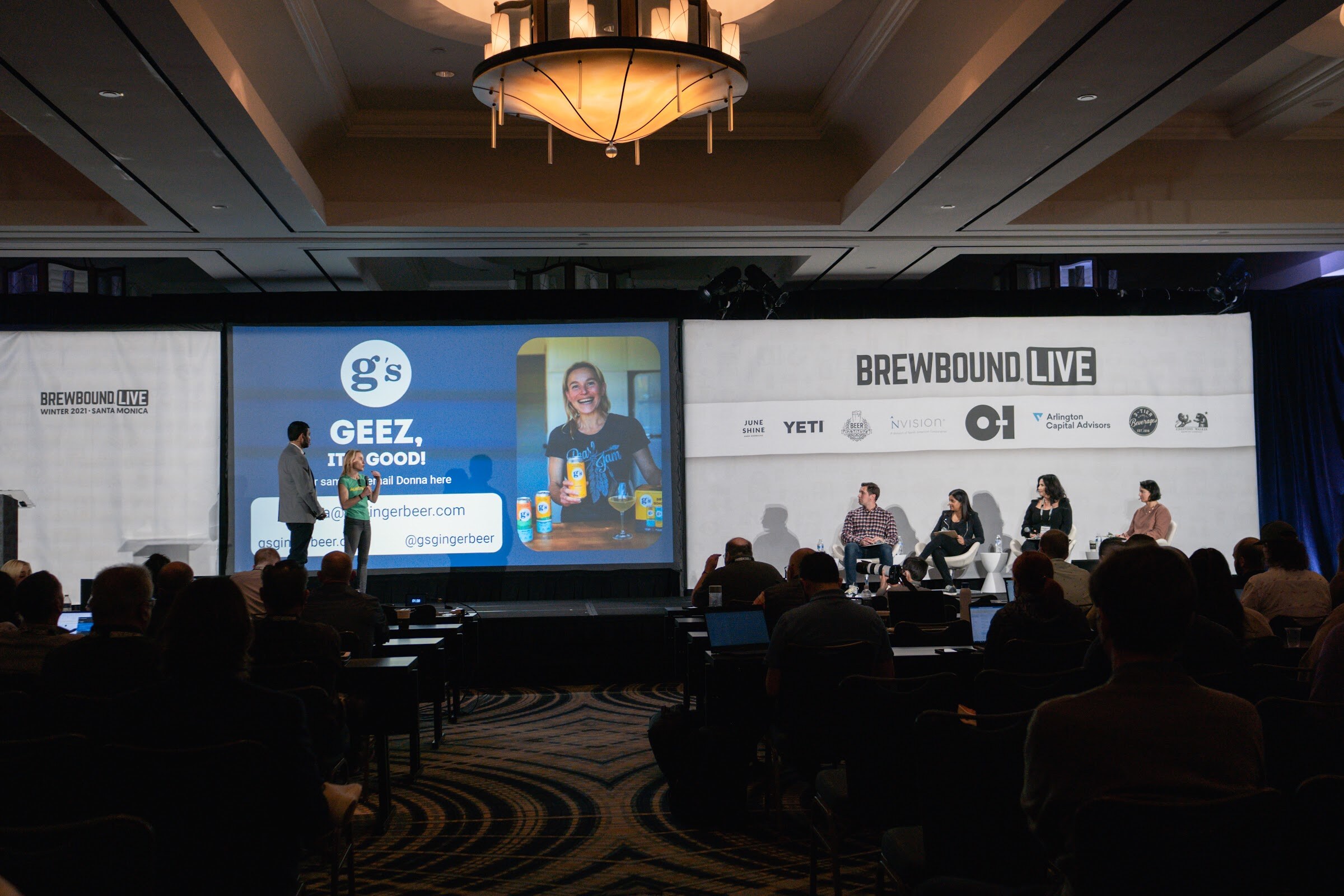 Wide view of a ballroom stage with a presenter in front of a G's Geez It's Good slide and four judges seated at right.