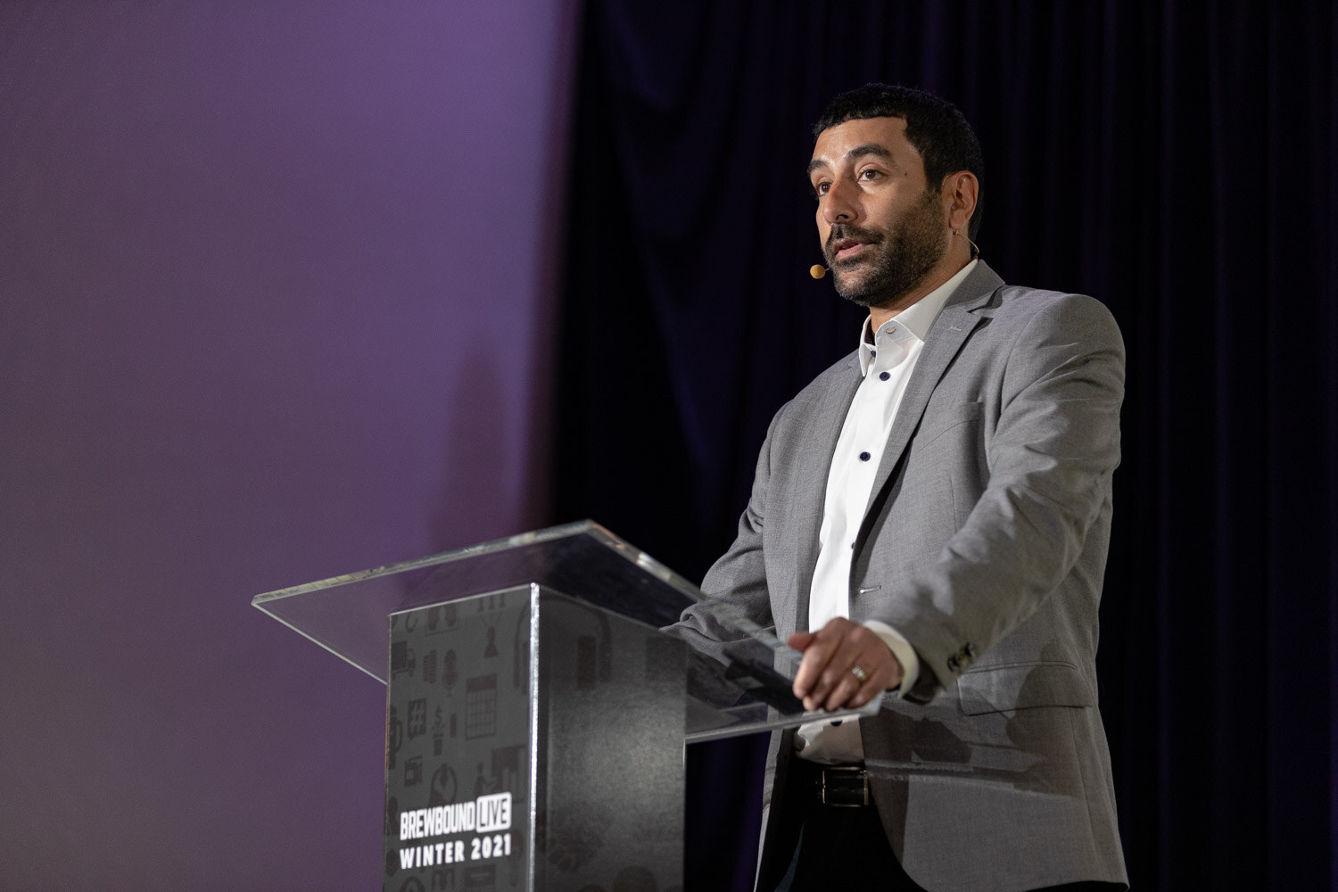 A man in a gray blazer speaks from a podium on a dark stage.