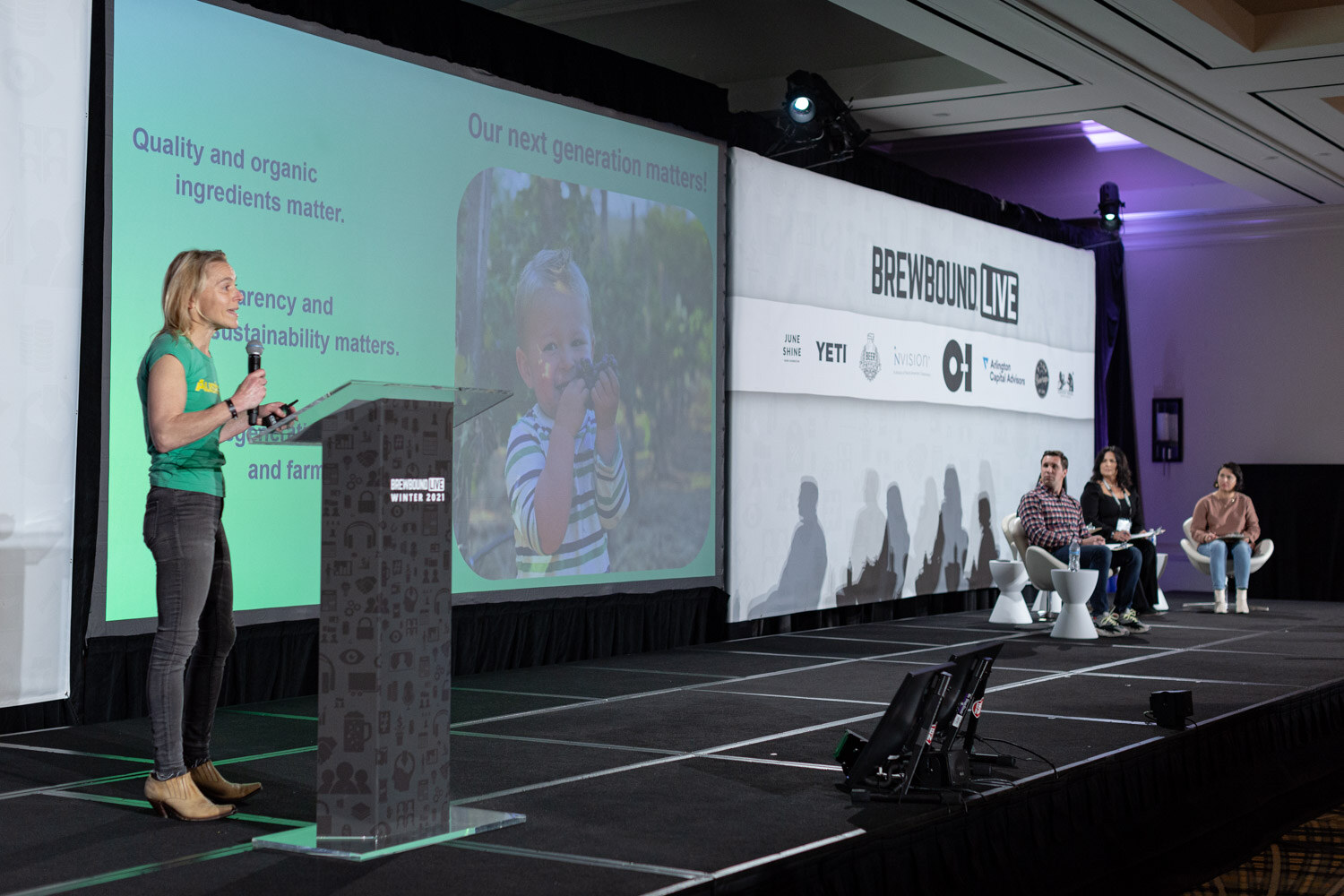 A woman in a green shirt stands at a podium beside a green slide about organic ingredients, with judges seated at right.