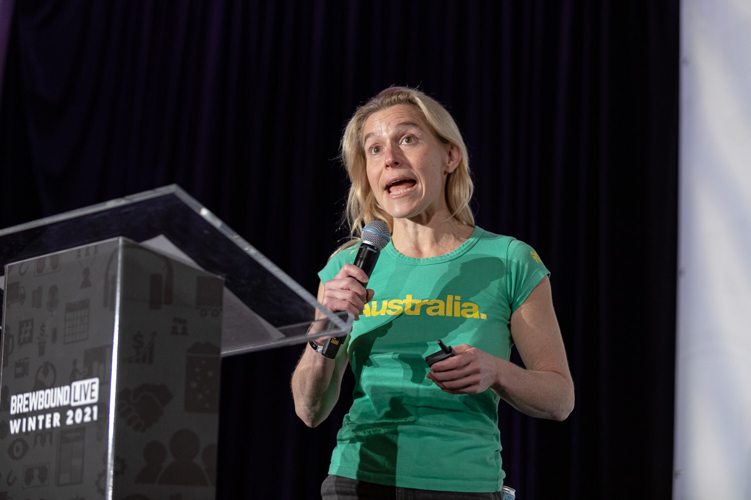 A woman in a green Australia t-shirt speaks into a microphone at a clear podium on a dark stage.