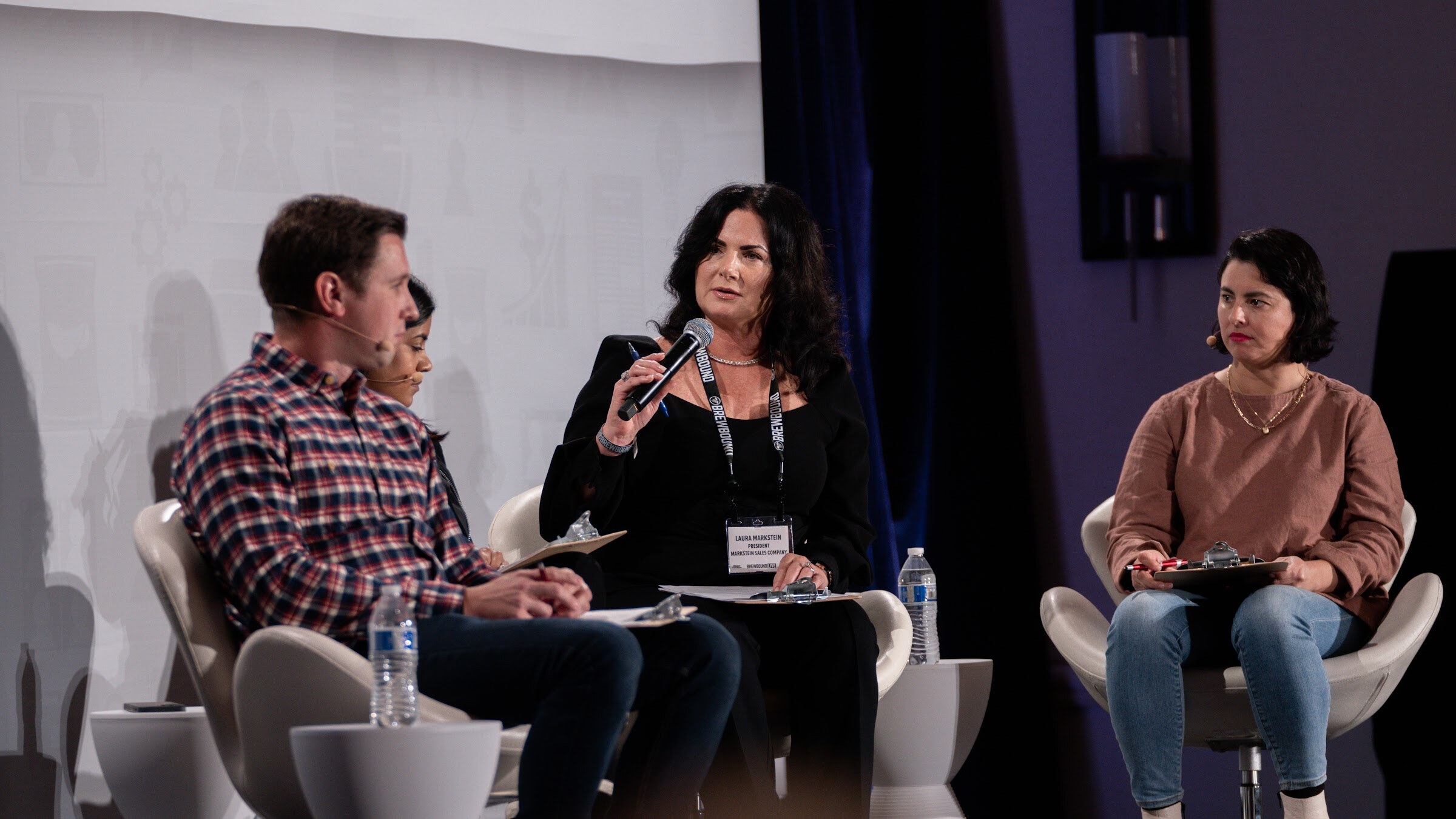 Three judges sit on stage, the woman in the middle speaking into a microphone while a man and a woman look on.