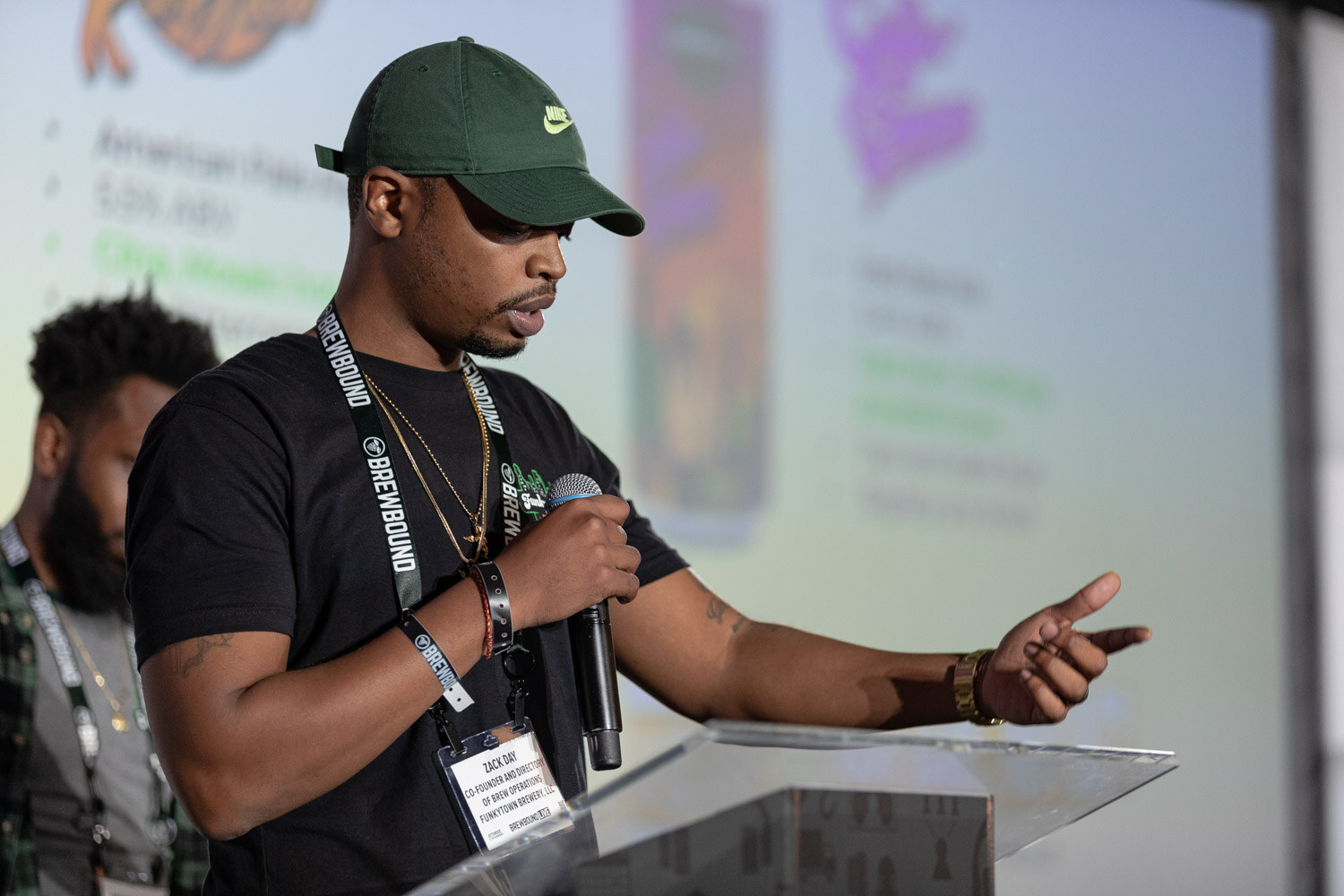 A man in a green cap holds a microphone and points while reading from a podium, with a teammate behind him.