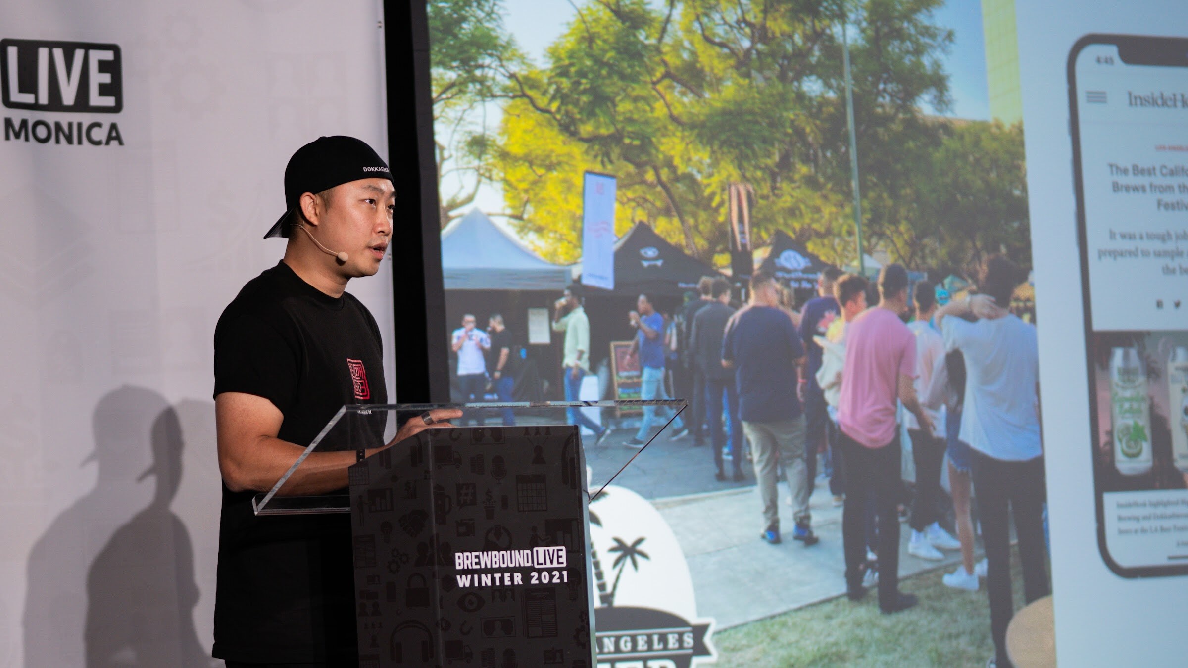 A man in a backwards cap speaks at a podium beside a slide showing a crowd at an outdoor beer festival.