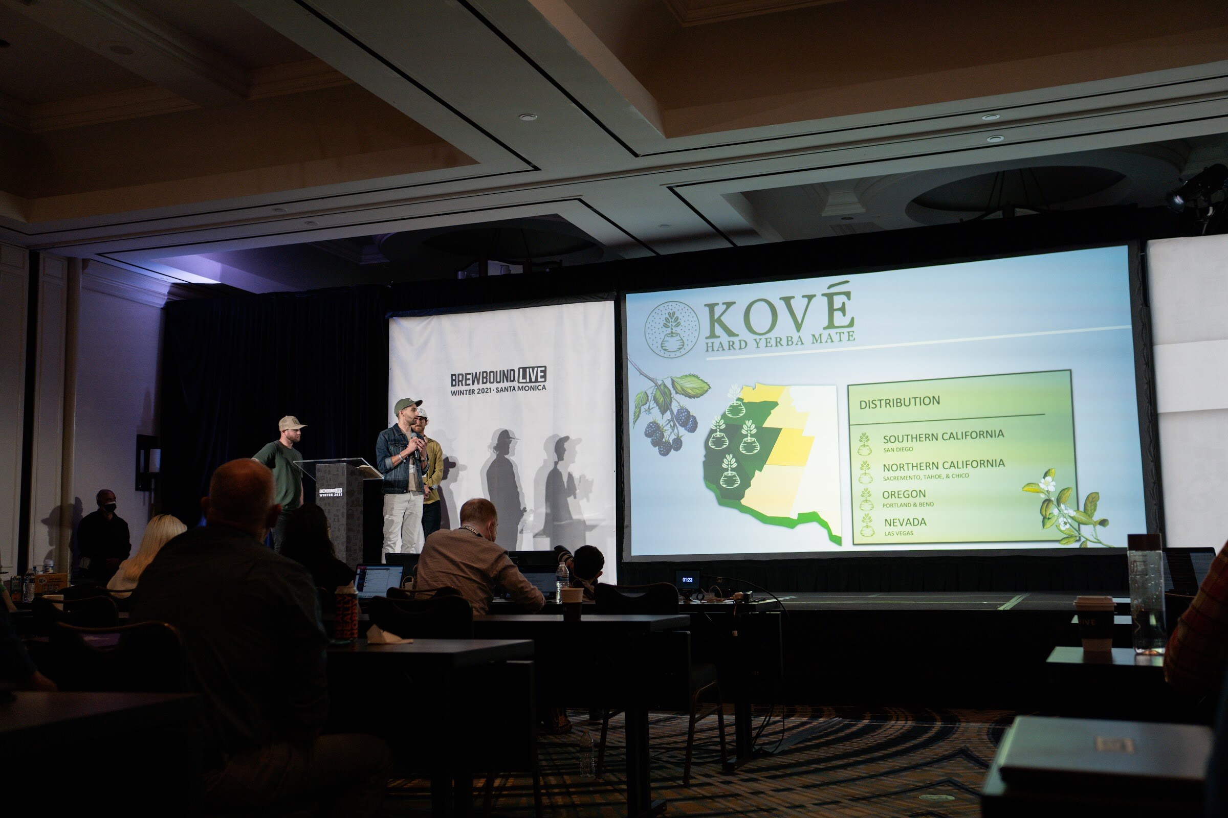 Wide view of a ballroom stage with two presenters at a podium and a Kove Hard Yerba Mate distribution slide on screen.