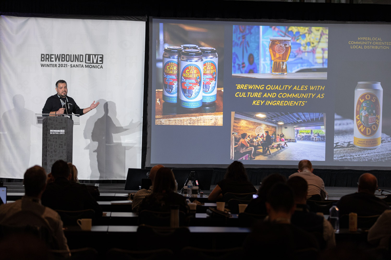 Wide view of a ballroom with a man at a podium beside a screen showing Los Car Mendoza cans and the quote Brewing quality ales with culture and community as key ingredients.