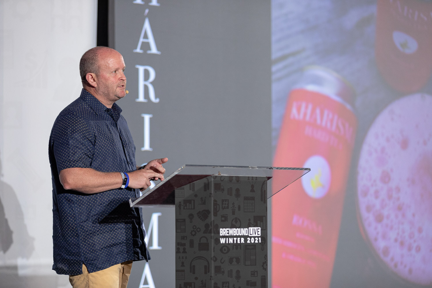 A man in a dark patterned shirt speaks at a podium in front of a large slide showing a Kharisma can.