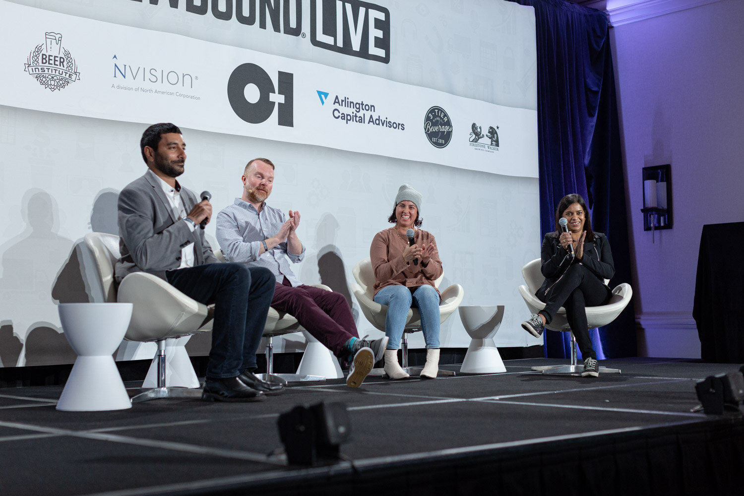 Four people sit on stage in front of a Brewbound Live banner with sponsor logos, clapping and holding microphones.