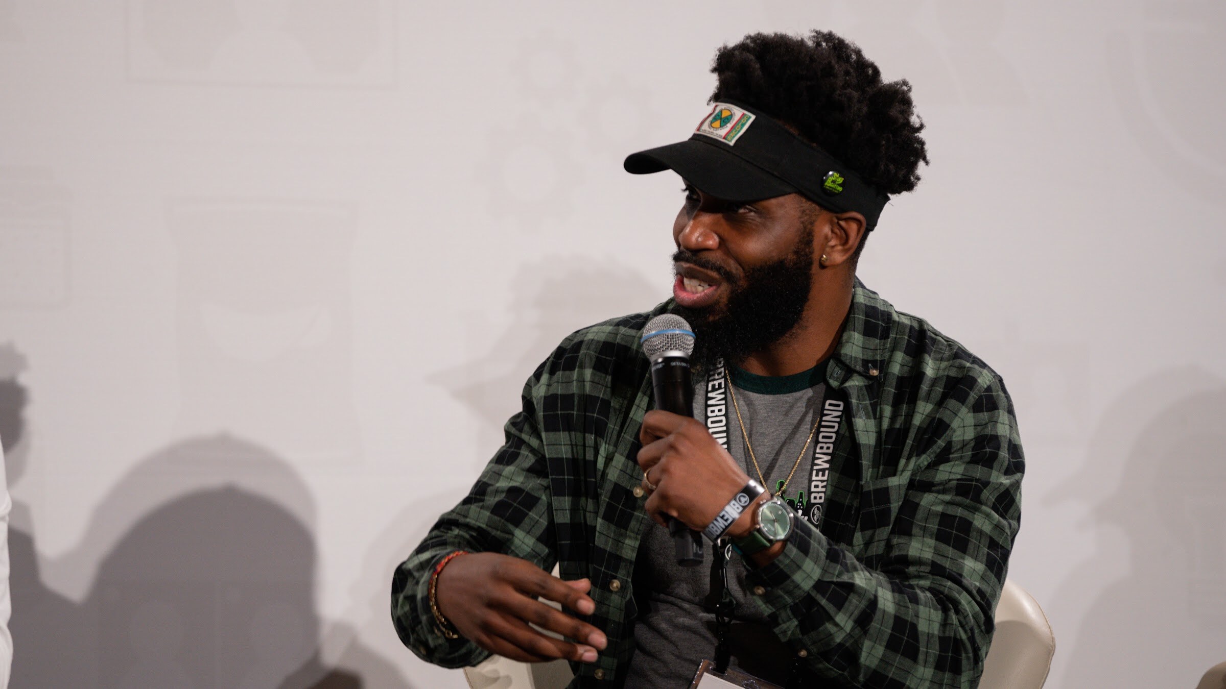 A bearded man in a visor and green plaid shirt speaks into a microphone while seated.