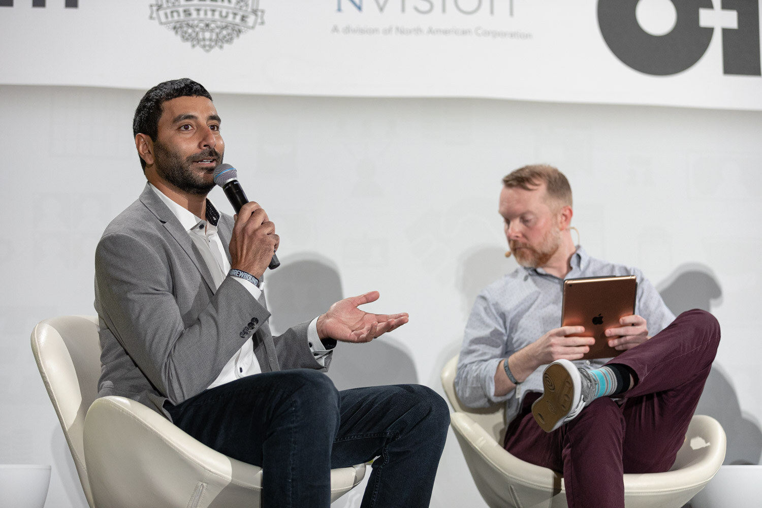 A man in a gray blazer holds a microphone and gestures beside a bearded man in light blue holding an iPad.