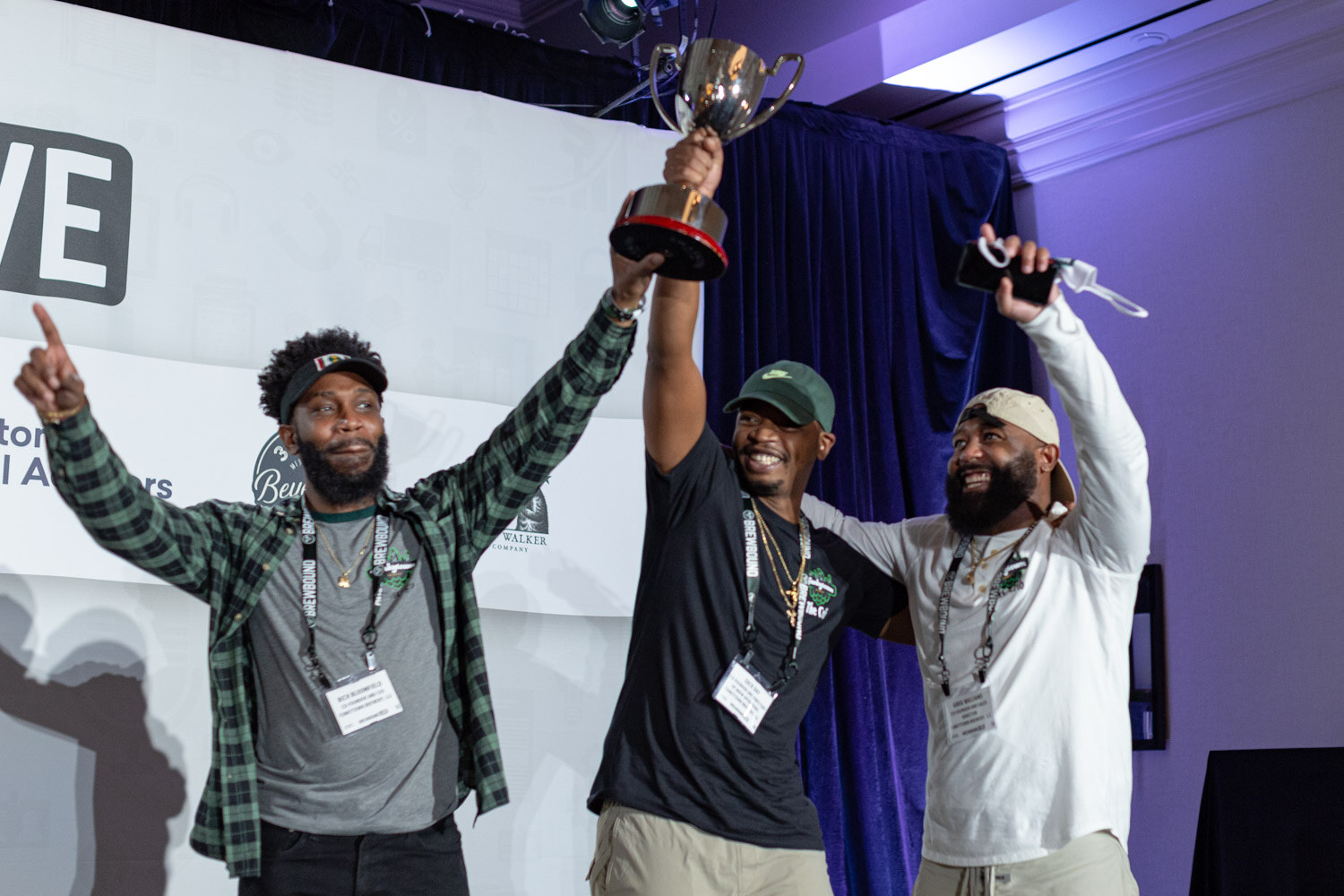 Three men with arms raised on stage, one holding a silver trophy cup over his head, with the Brewbound Live backdrop behind them.