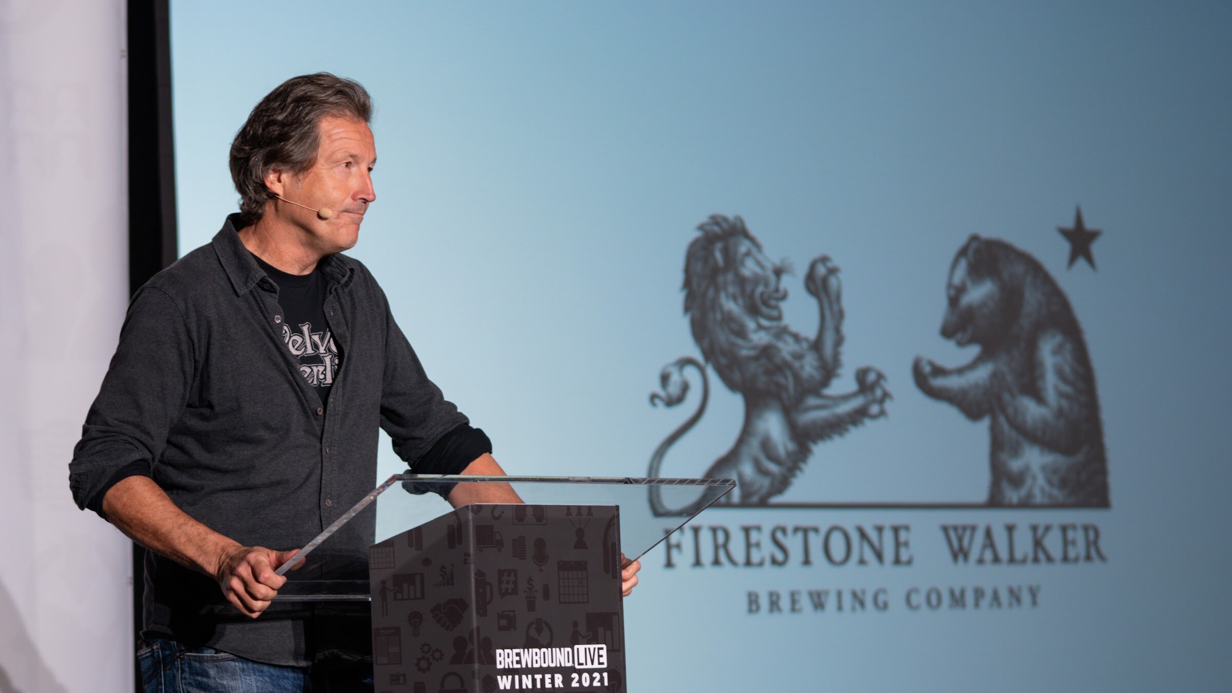 A man in a dark shirt stands at a podium in front of a screen showing the Firestone Walker lion and bear logo.