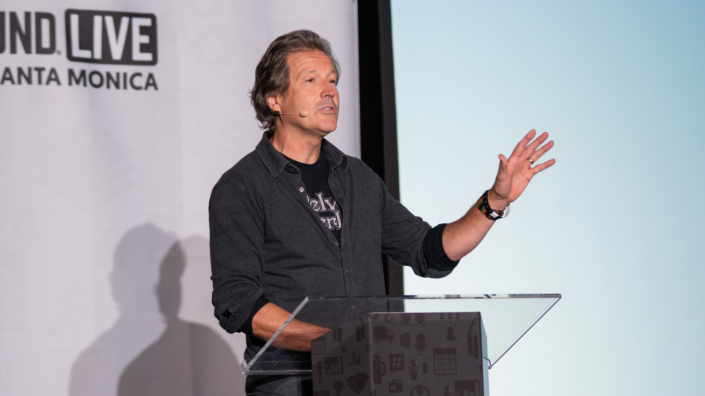 A man in a dark button-up shirt gestures with one hand beside a clear podium, with a Brewbound Live banner behind him.
