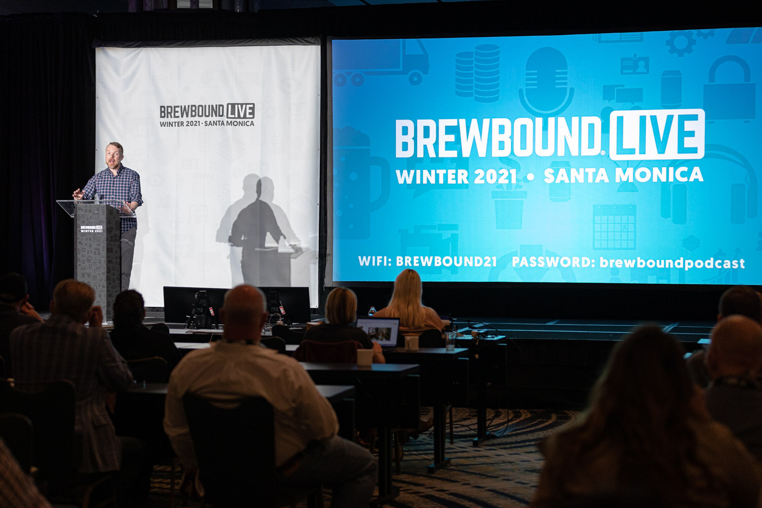 Wide view of a ballroom with a man at a podium, a Brewbound Live Winter 2021 Santa Monica screen and attendees seated at tables.