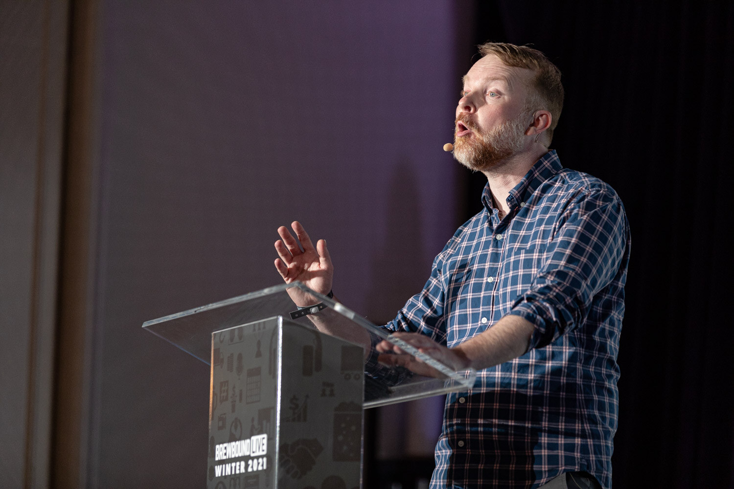 A man in a plaid shirt speaks animatedly at a clear podium on a dark stage.