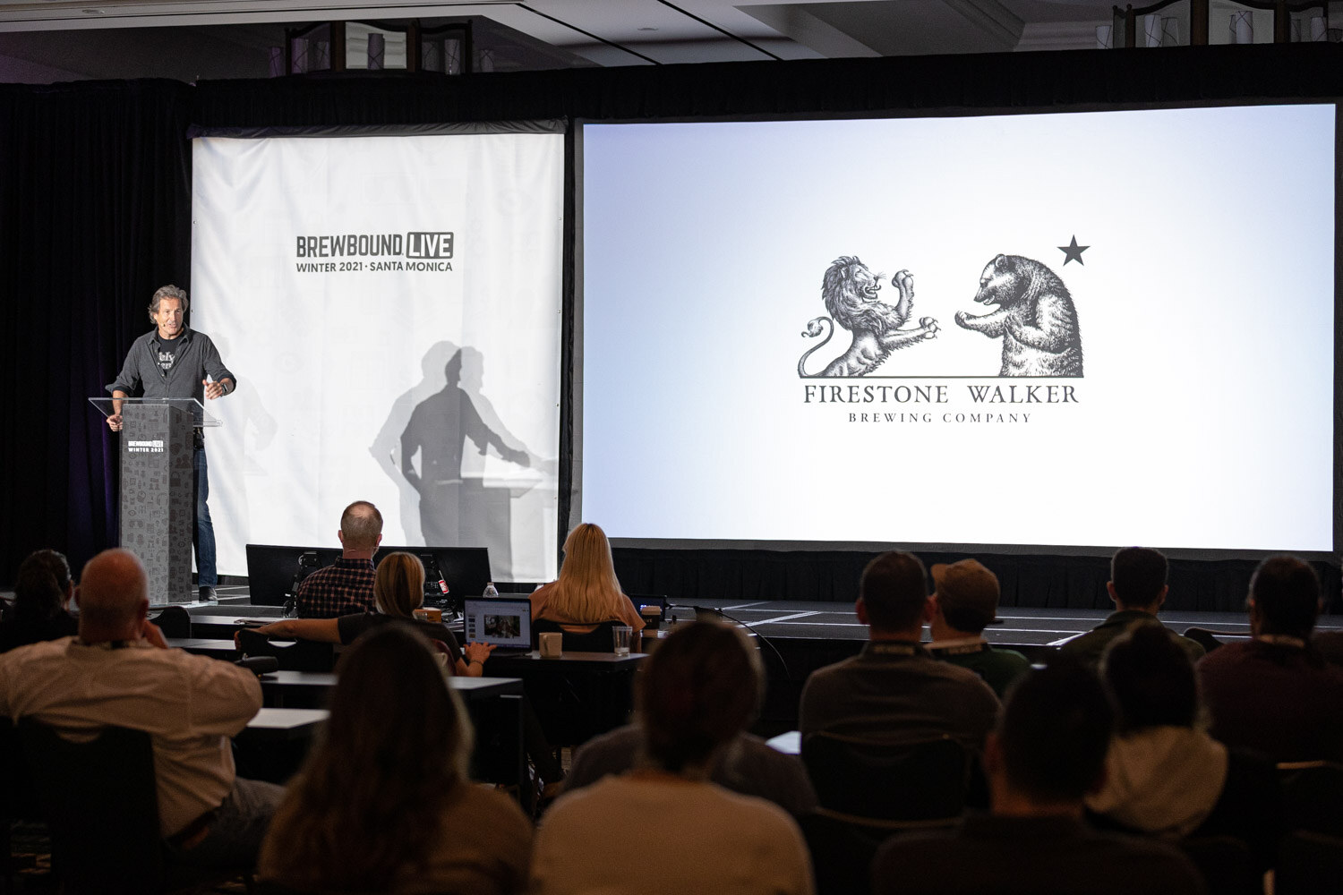 Wide view of a ballroom stage with a man at a podium, a Firestone Walker logo on the screen and attendees seated in the foreground.