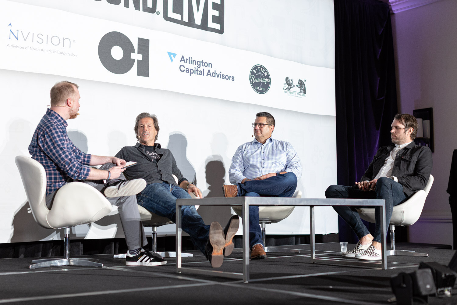Four men sit in white chairs around a low table on stage in front of Brewbound Live sponsor logos.