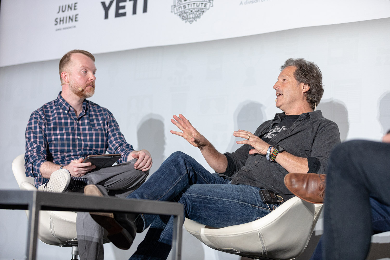 Two men seated in white chairs talk on stage, one holding a tablet and the other gesturing with both hands.