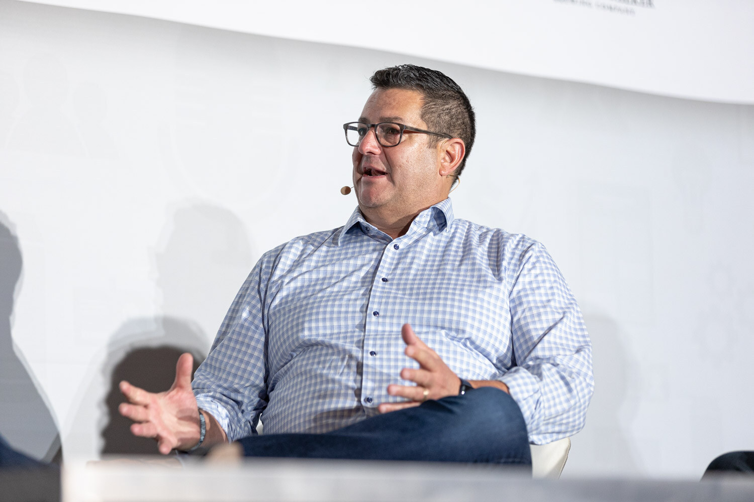 A man in glasses and a light checked shirt gestures with both hands while seated on stage.