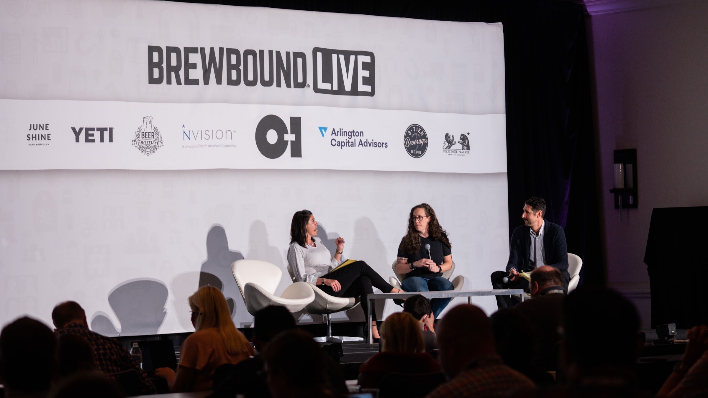 Three people sit in white chairs on a stage in front of a Brewbound Live backdrop with sponsor logos, a man at right speaking into a microphone.