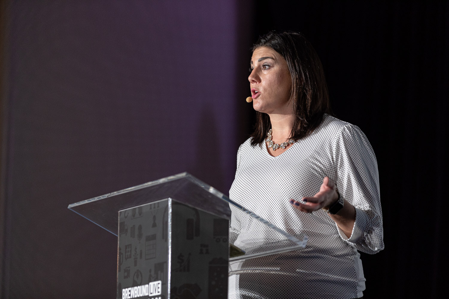 A woman in a white patterned top speaks at a clear podium on a dark stage.