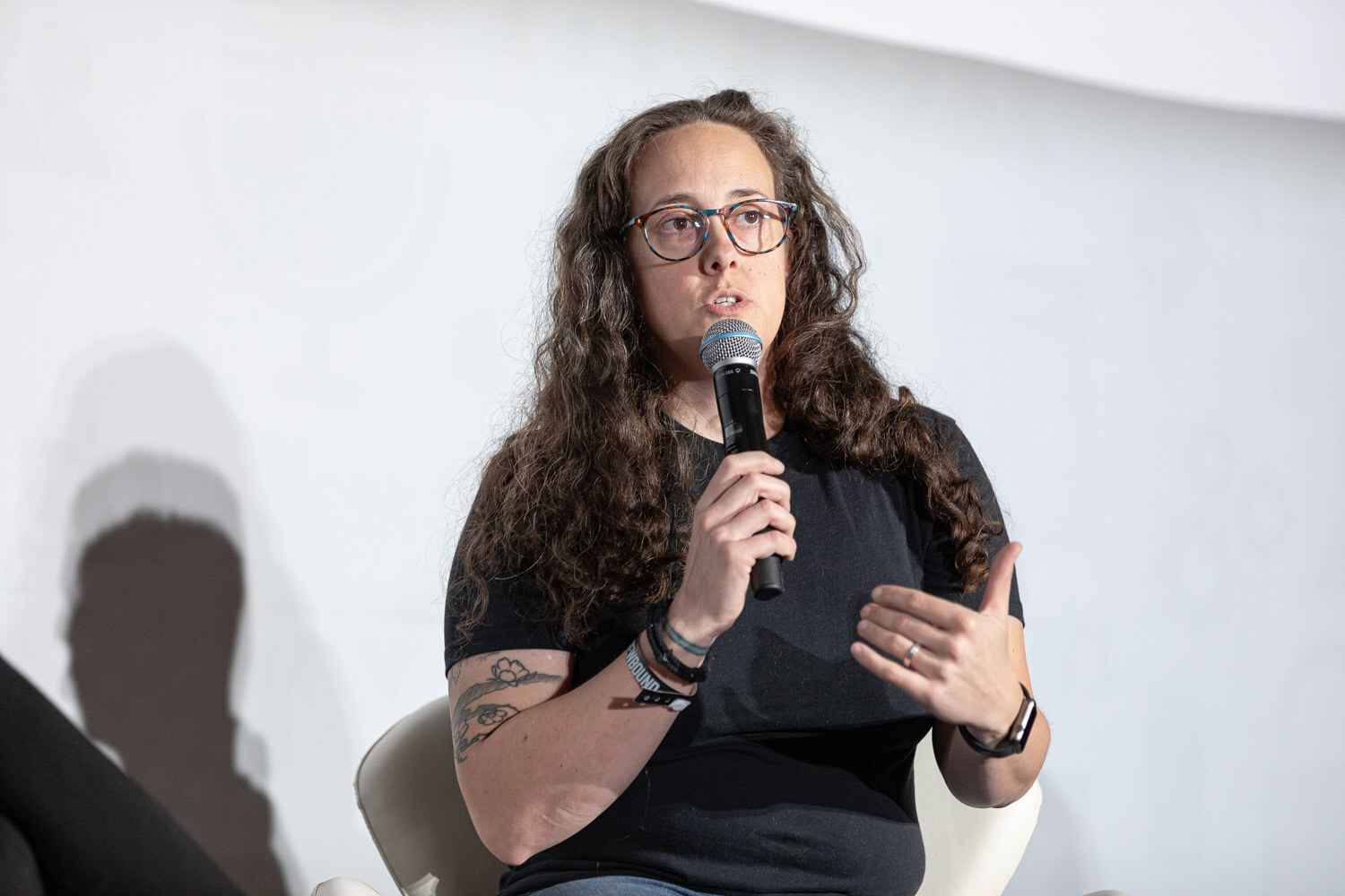 A woman with long curly hair and glasses holds a microphone and gestures while seated.