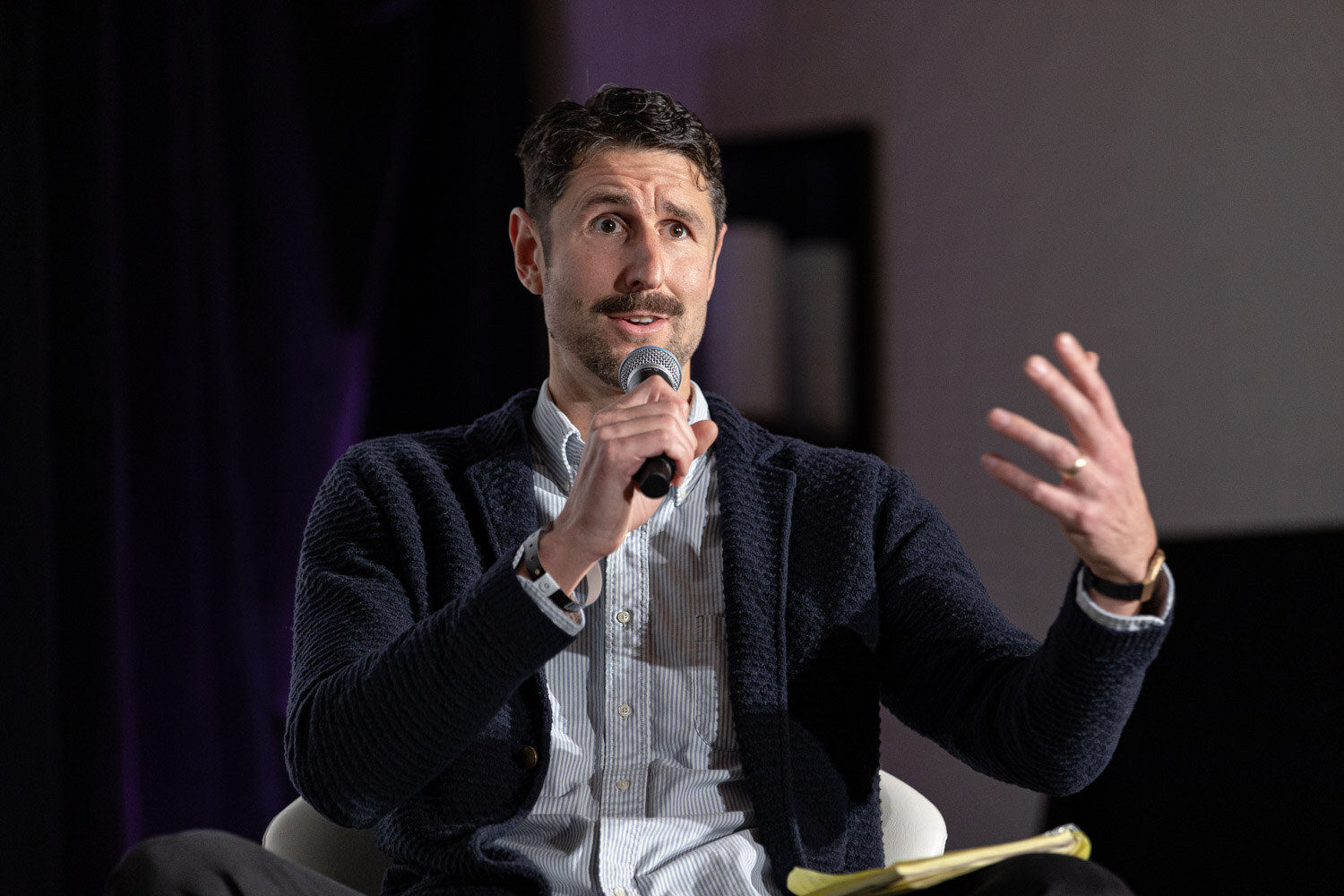 A man with a mustache in a navy cardigan holds a microphone and raises one hand while holding a yellow notepad.