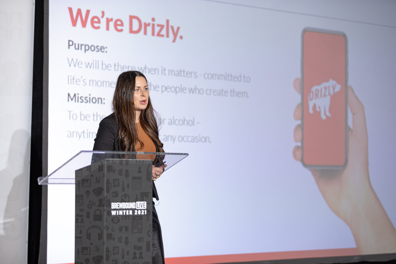 A woman stands at a podium in front of a slide reading We're Drizly with a phone showing the Drizly app.