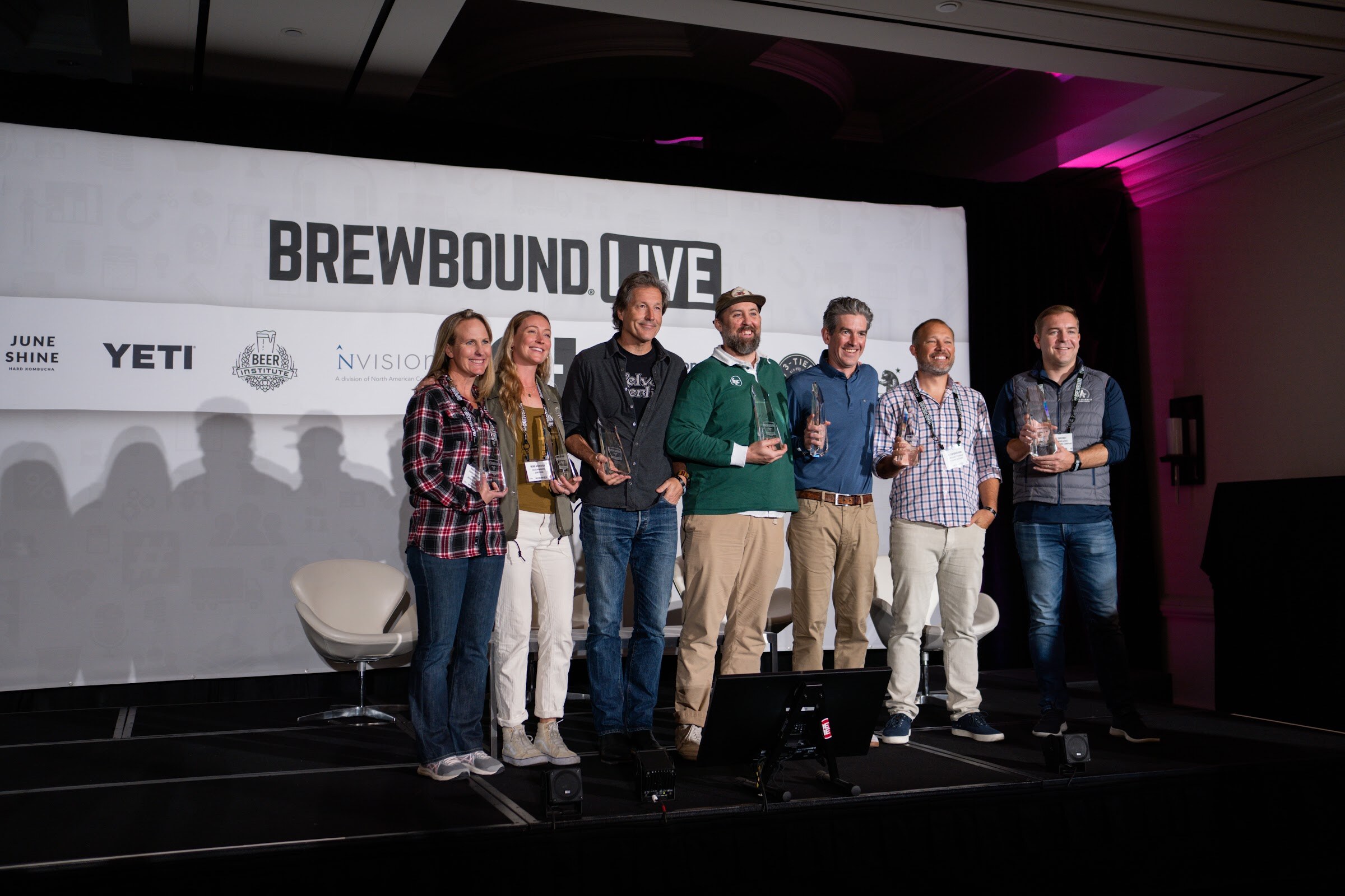 Seven people stand in a row on a stage holding glass trophies in front of a Brewbound Live banner.