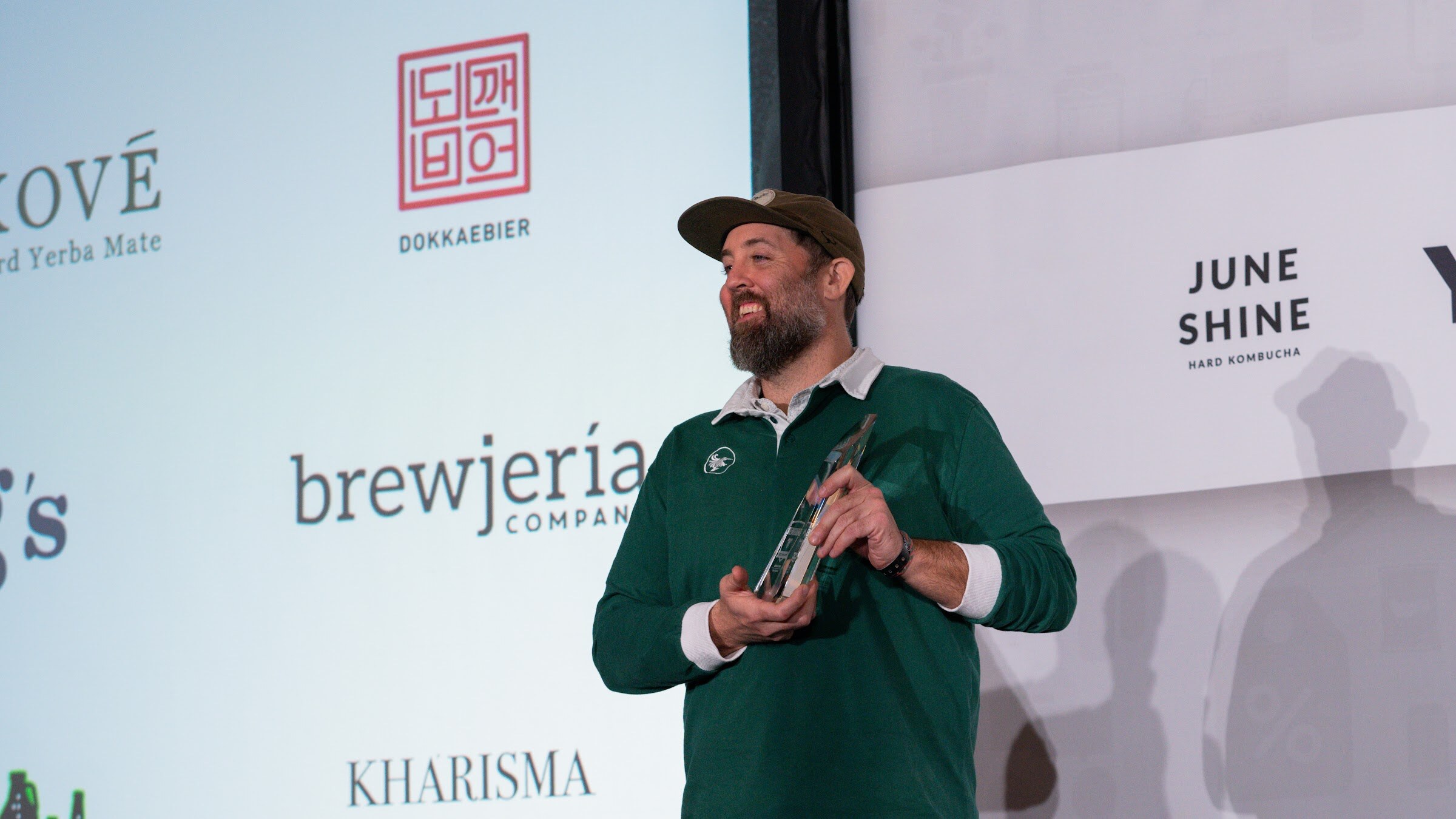 A bearded man in a green rugby shirt holds a glass trophy in front of a screen showing company logos including Kove, Dokkaebier, brewjeria and Kharisma.