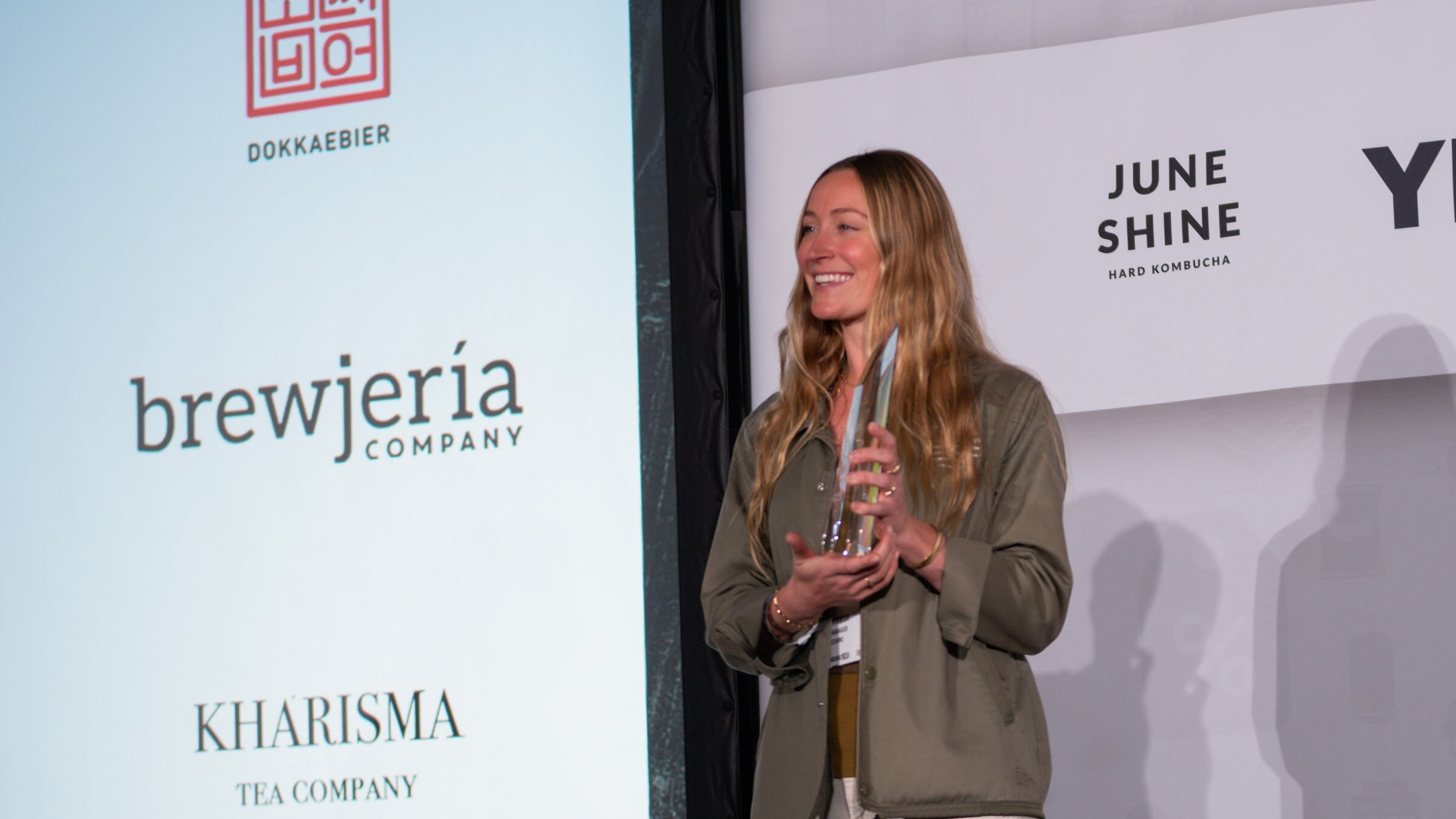 A woman in an olive jacket stands on stage holding a glass trophy in front of a screen with Dokkaebier, brewjeria and Kharisma logos.