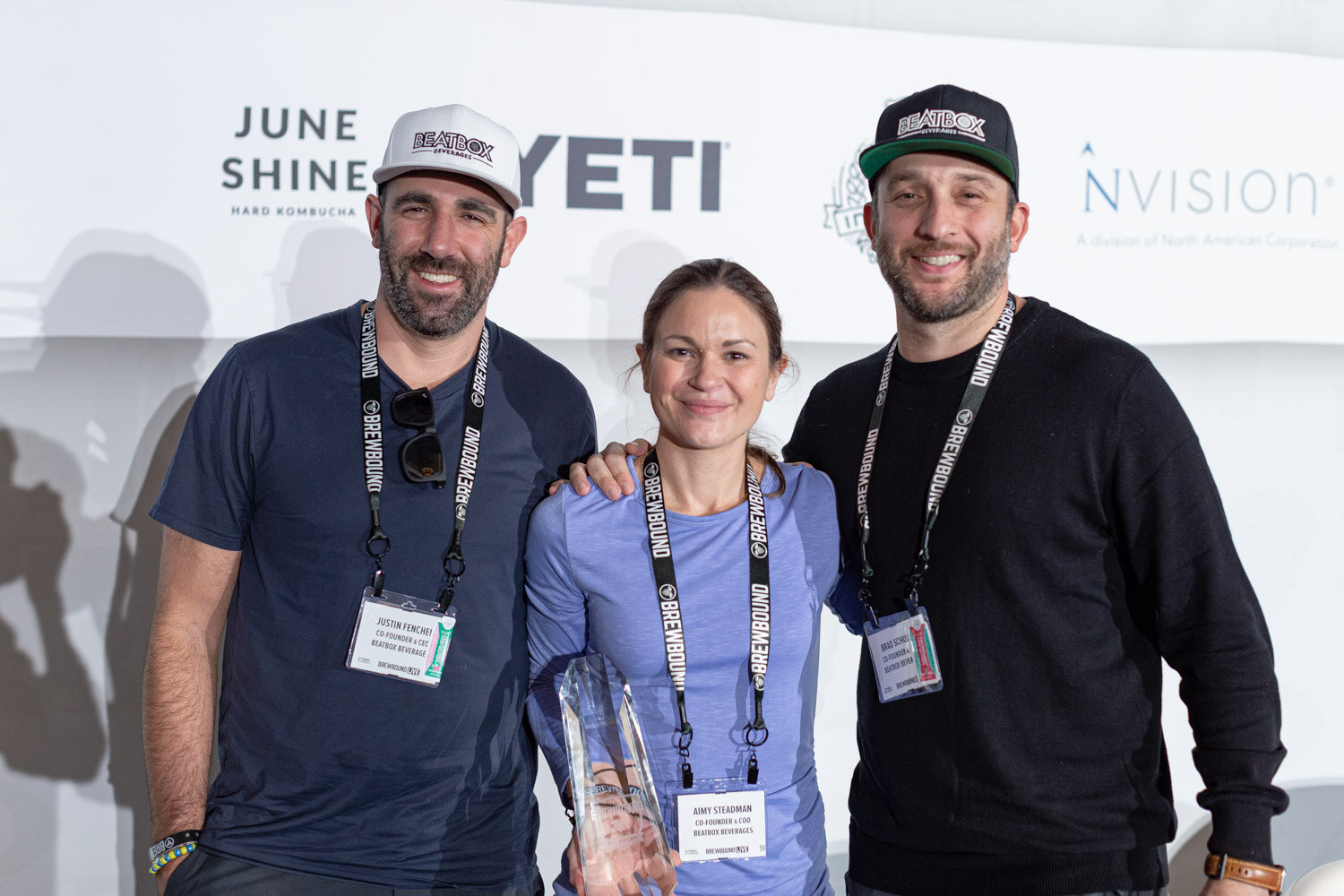 Two men and a woman in lanyards pose together in front of a sponsor backdrop; the woman holds a glass trophy.