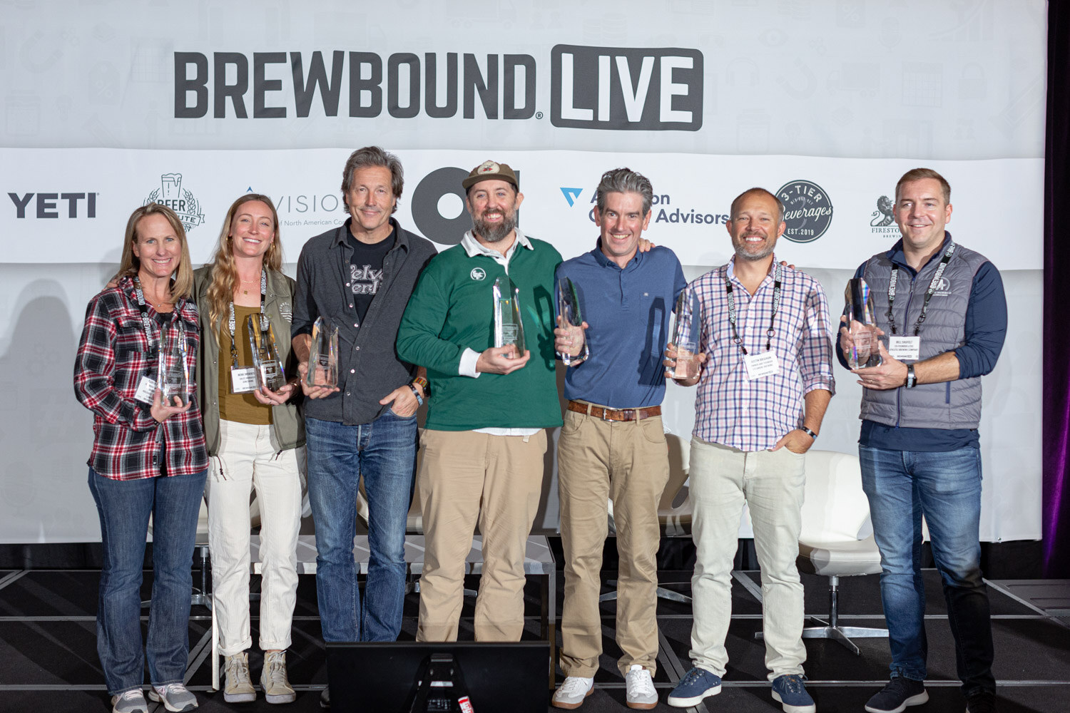 Seven people stand in a line holding glass trophies in front of a Brewbound Live backdrop.