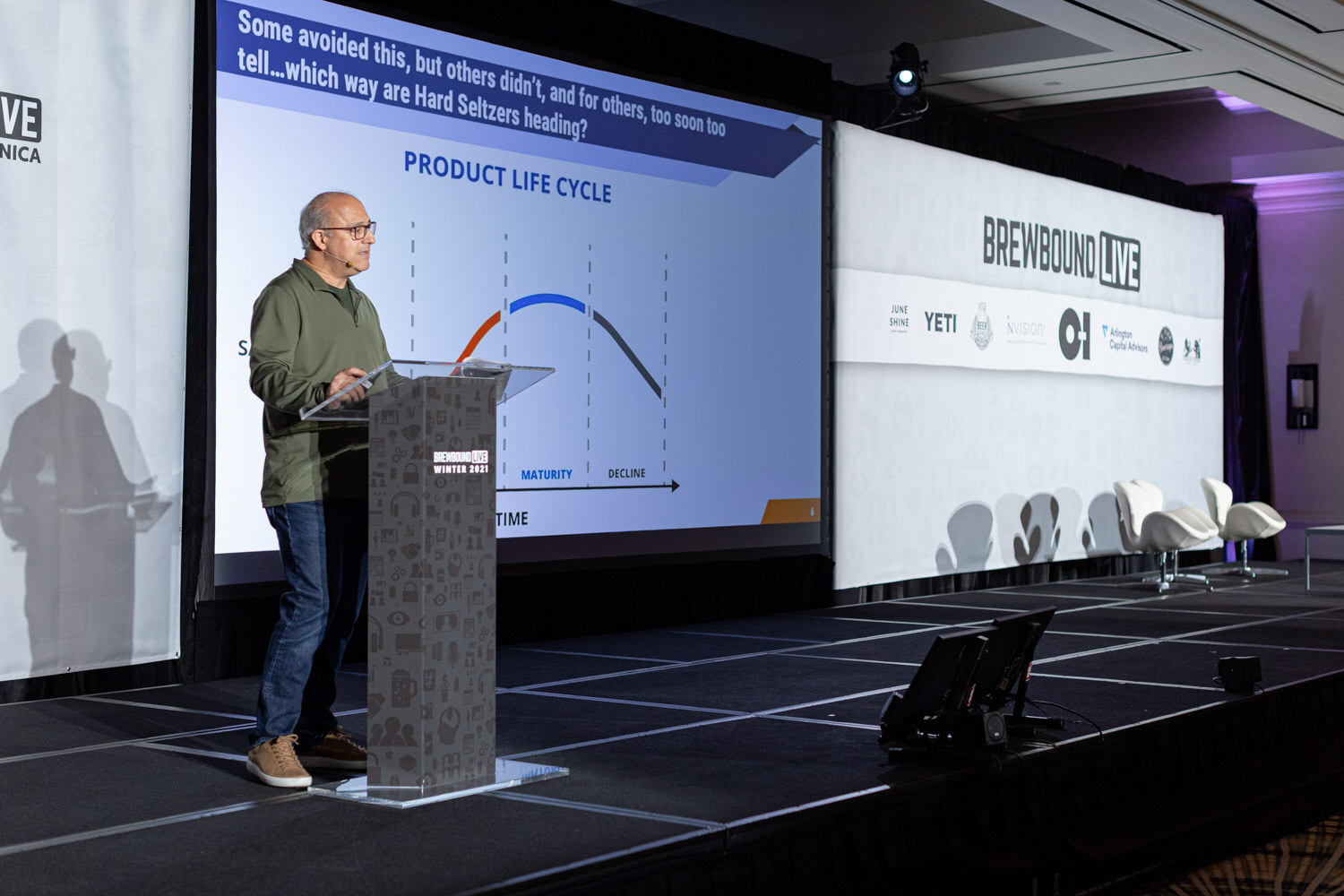 A man in a green pullover stands at a podium beside a slide titled Product Life Cycle with a curve rising to maturity and decline.