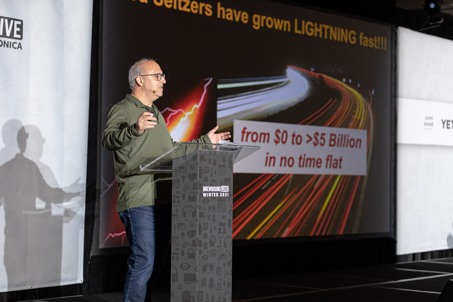 A man in glasses gestures at a podium in front of a slide reading Seltzers have grown lightning fast, from $0 to $5 billion in no time flat.