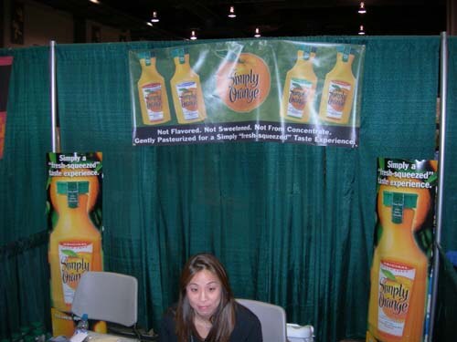 A woman with long dark hair sits at a table in front of a green curtain with Simply Orange banners showing orange juice bottles.