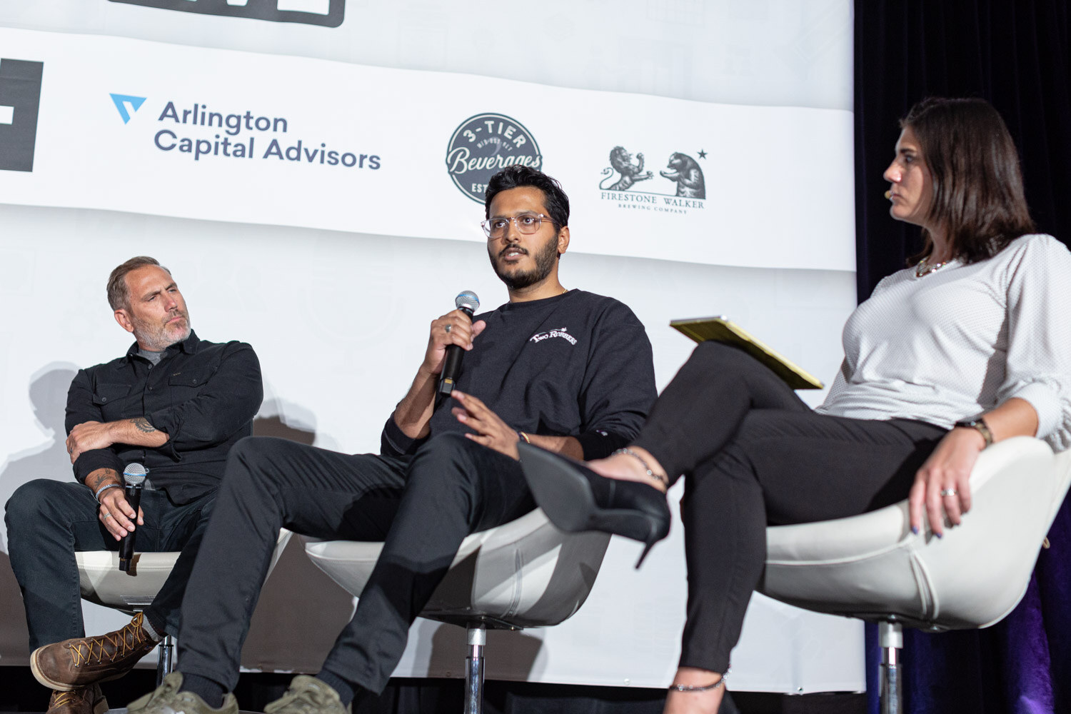 A man in a black sweatshirt speaks into a microphone between a man with folded arms and a woman with a yellow notepad.