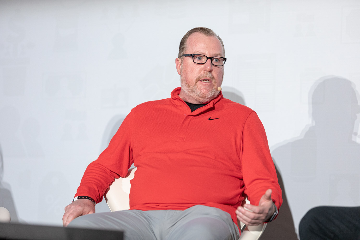 A man in glasses and a red pullover speaks while seated on stage.