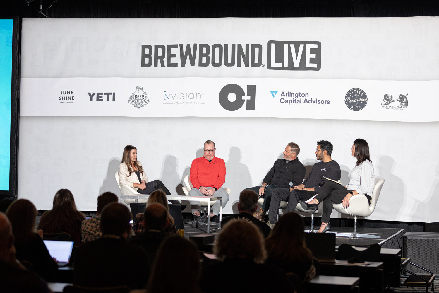 Five people sit in a row on a stage under a Brewbound Live banner with sponsor logos while attendees with laptops watch.