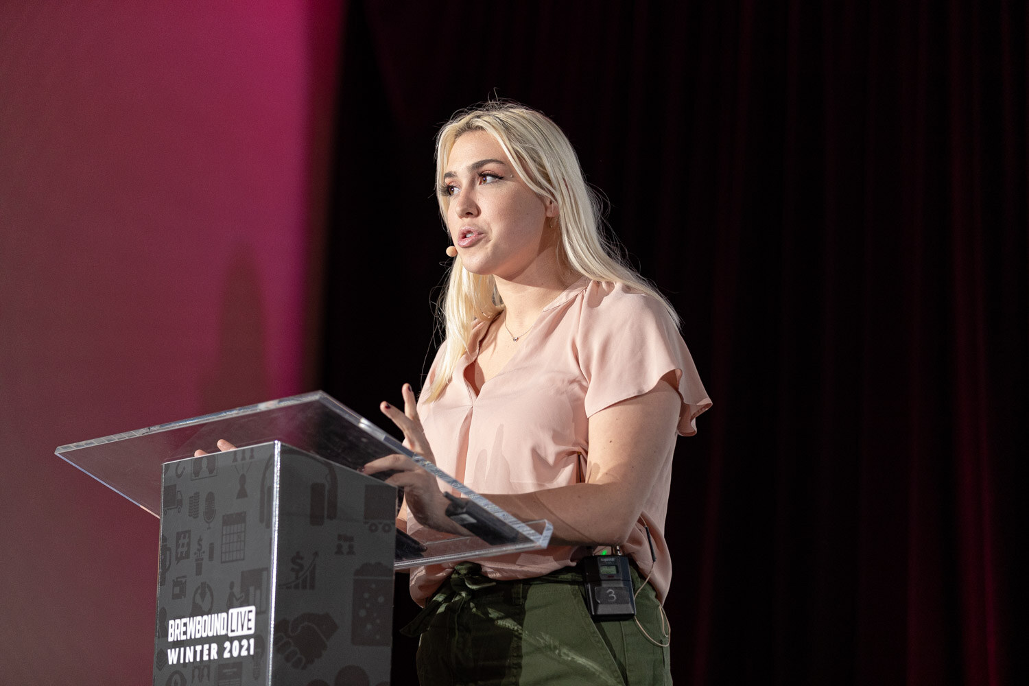 A woman with long blonde hair in a pale pink top speaks at a clear podium.