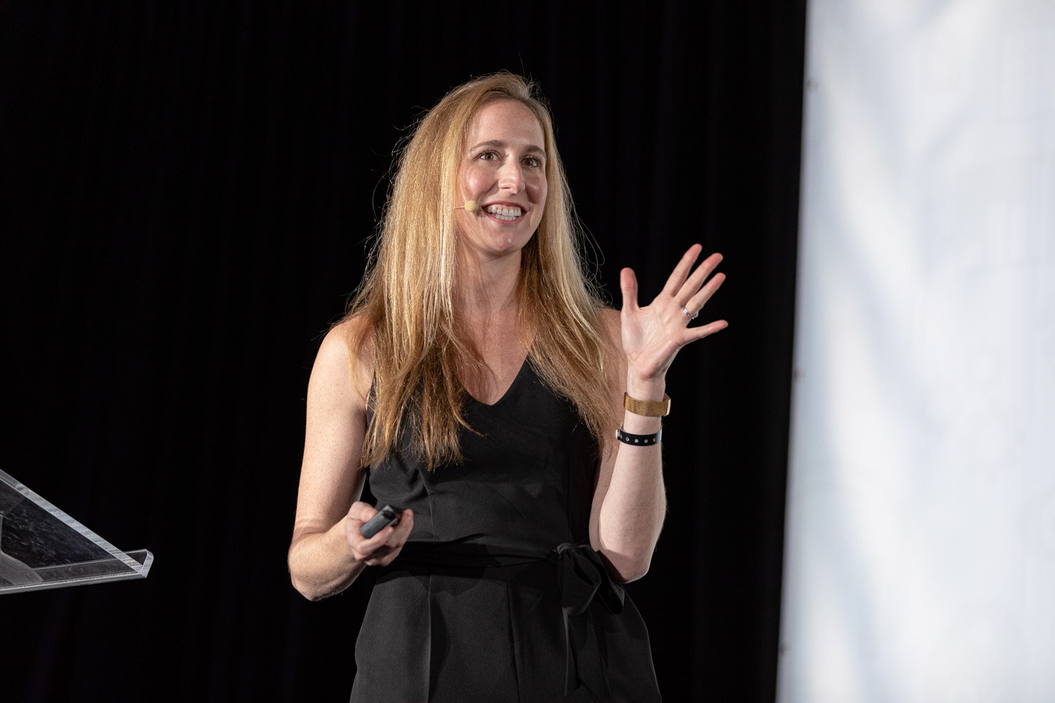 A woman in a black sleeveless outfit smiles with one hand raised while holding a clicker.