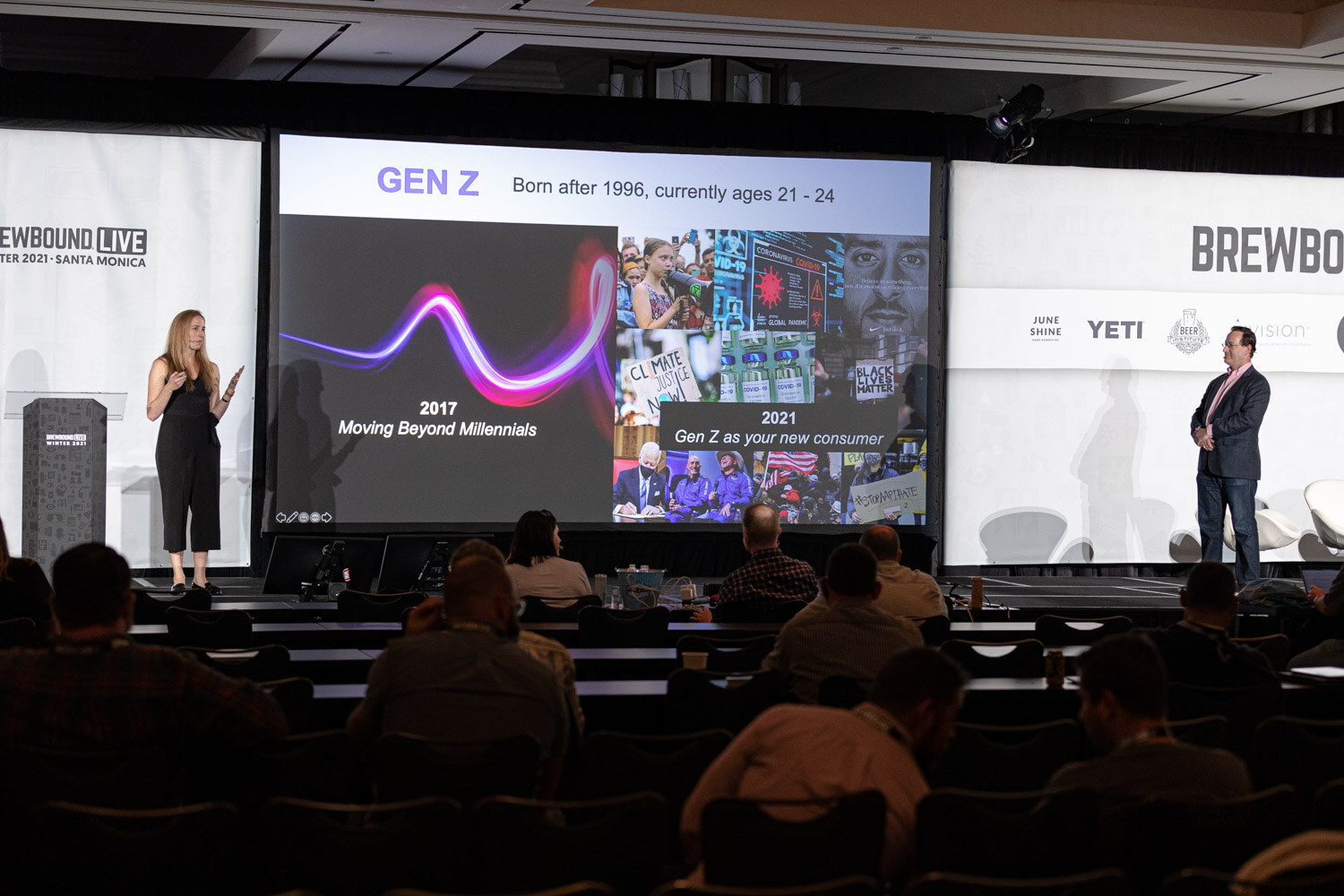 A woman stands at left and a man at right on a stage beside a large screen reading Gen Z, born after 1996, currently ages 21 to 24.
