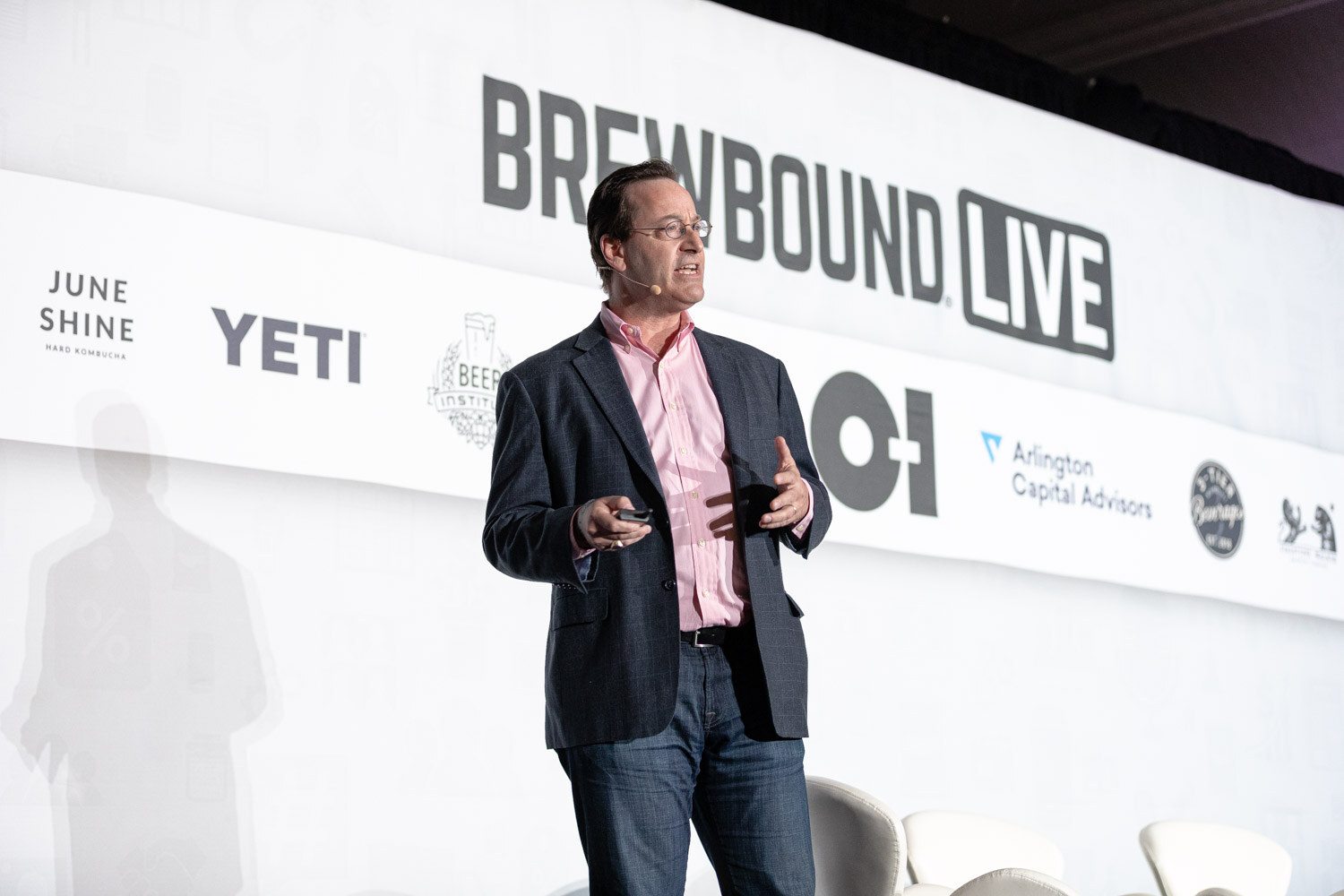 A man in glasses, a blazer and a pink shirt walks in front of a Brewbound Live banner with sponsor logos.