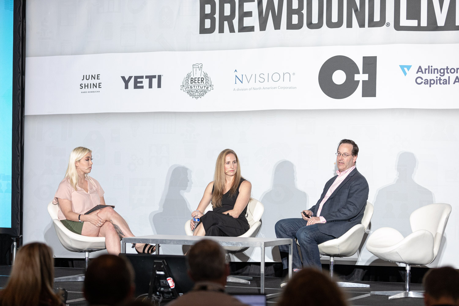 Three people sit on white chairs on stage in front of a Brewbound Live banner while audience heads fill the foreground.