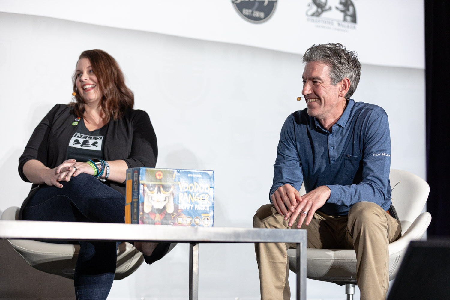 A woman in a Bell's shirt and a man in a blue pullover sit on stage laughing, with a box of beer on a low table between them.