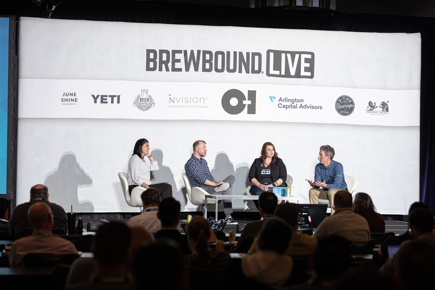 Four people sit on stage in front of a Brewbound Live backdrop with sponsor logos while attendees watch from the foreground.