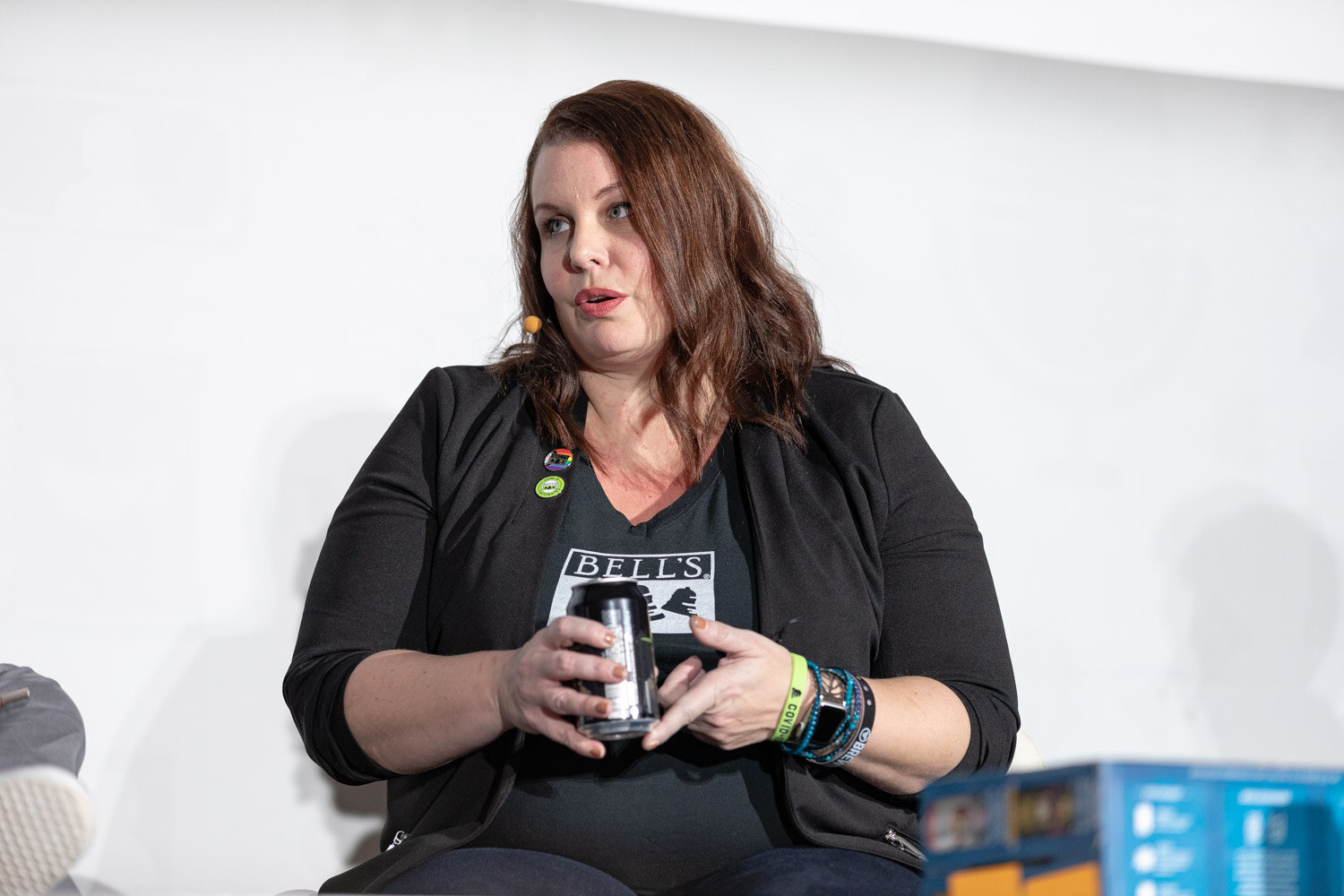A woman in a black Bell's shirt holds a can in both hands and speaks while seated on stage.