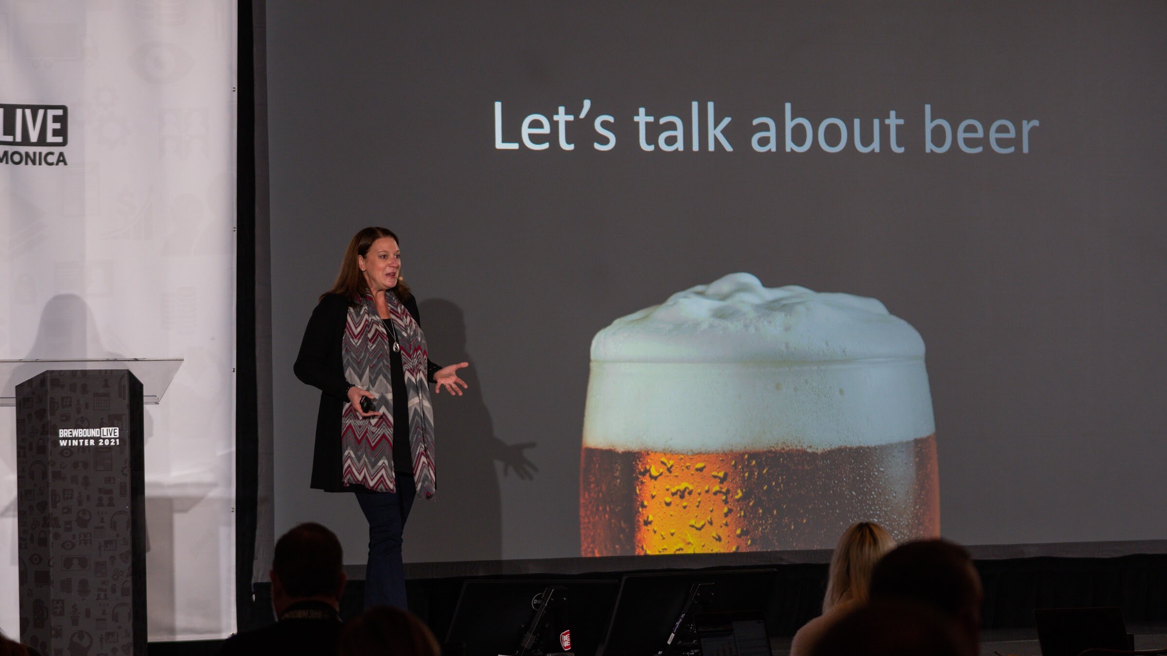 A woman in a scarf walks the stage beside a podium in front of a large slide reading Let's talk about beer with a glass of beer.