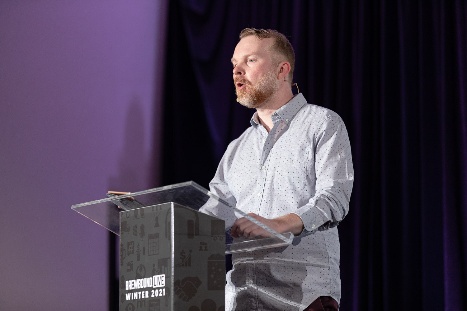 A bearded man in a light dotted shirt speaks at a clear podium in front of a dark purple curtain.